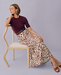 A woman in a patterned skirt and maroon top sits on a chair against a lavender background. carousel Product Image 4