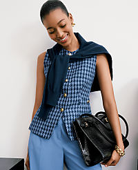 A smiling woman in a houndstooth vest and blue pants holds a black handbag against a plain white background. carousel Product Image 3