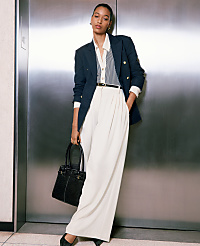A woman in a navy blazer and white pants stands in front of a brushed metal elevator door, holding a black handbag. carousel Product Image 5