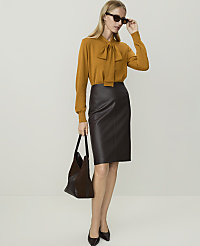 A woman in a mustard-colored bow-neck sweater and a dark brown leather pencil skirt stands with a dark handbag against a plain white background. carousel Product Image 1