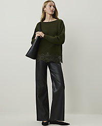 A woman in a green sweater and black leather pants stands against a plain white background, holding a black tote bag. carousel Product Image 1