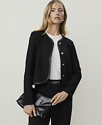 A woman in a black jacket and pants holds a black clutch in front of a plain background. carousel Product Image 1