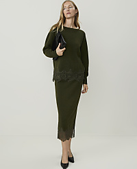A woman in a green sweater and skirt with lace trim stands against a plain white background, holding a black clutch. carousel Product Image 4