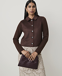 A woman in a brown jacket and patterned skirt holds a woven clutch in front of a plain background. carousel Product Image 1