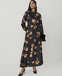 A woman in a floral dress stands against a plain white background, holding a black clutch. carousel Product Image 3
