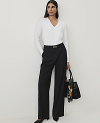 A woman in a white v-neck sweater and black wide-leg pants stands against a plain white background, holding a black tote bag. carousel Product Image 4