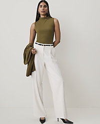 A woman in a green mock-neck top and white wide-leg pants stands against a plain, light background, holding a green jacket over her arm. carousel Product Image 4