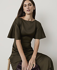 A woman in a flowing olive green dress sits on a stool in front of a plain white background. carousel Product Image 3