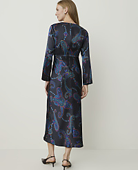 A woman in a long, dark, paisley-patterned dress stands against a plain, light-colored background. carousel Product Image 2