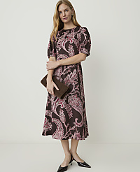 A woman in a brown paisley dress and black heels holds a brown clutch in front of a plain white background. carousel Product Image 1