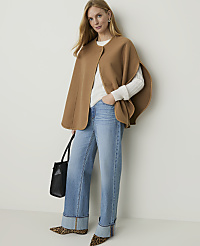 A woman in a camel cape, white sweater, and cuffed jeans stands with a black bag, against a plain background. carousel Product Image 1