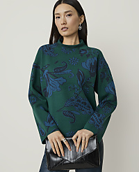 A woman in a green patterned sweater holds a black clutch in front of a plain background. carousel Product Image 1