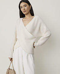 A woman in a cream wrap sweater and white pants stands against a plain, light background, holding a woven bag. carousel Product Image 1