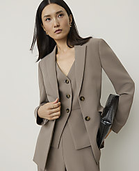 A woman in a taupe suit and holding a black clutch stands against a plain, light background. carousel Product Image 1