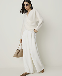 A woman in a cream wrap sweater and wide-leg pants poses against a plain, light background, holding a woven handbag. carousel Product Image 1