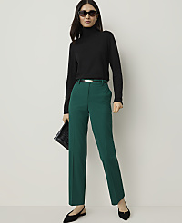 A woman in a black turtleneck and green pants stands against a plain white background, holding a black clutch. carousel Product Image 1