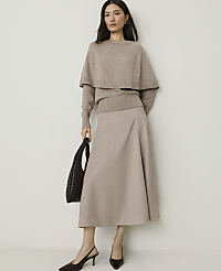 A woman in a beige sweater and skirt stands against a plain white background, holding a dark brown handbag. carousel Product Image 3