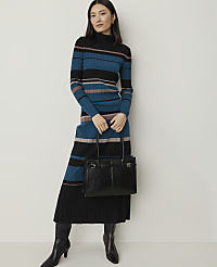 A woman in a striped turtleneck dress and black boots holds a black crocodile-embossed handbag against a plain white background.