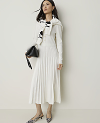 A woman in a white pleated dress and a floral-patterned sweater draped over her shoulders holds a black clutch while standing against a plain white background. carousel Product Image 1