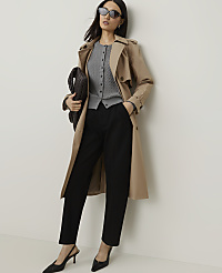 A woman in a trench coat, cardigan, and black pants poses against a plain white wall, holding a dark clutch. carousel Product Image 1