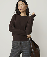A woman in a brown sweater and plaid pants stands against a plain background, holding a brown bag. carousel Product Image 1
