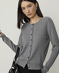 A woman in a grey cable-knit cardigan stands against a plain, light background, with a portion of a striped bag visible in the foreground. carousel Product Image 1