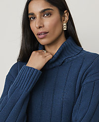 A woman with dark hair and a teal turtleneck sweater is in the foreground, while a plain, light-colored background is visible behind her. carousel Product Image 1