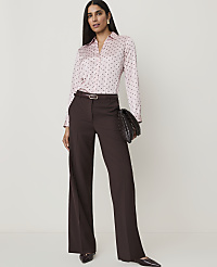 A woman in a pink polka dot blouse and brown wide-leg pants stands against a plain white background, holding a woven clutch. carousel Product Image 1