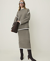 A woman in a taupe knit sweater and skirt with white trim, holding a brown suede tote bag, stands against a plain, light-colored background.
