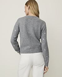 A woman in a grey sweater with embroidered details on the sleeves and white pants stands against a plain, light grey background. carousel Product Image 2