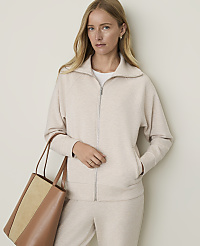 A woman in a cream-colored zip-up jacket and matching pants stands with a brown and tan tote bag in the foreground against a plain, light-colored background.