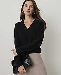 A woman in a black v-neck sweater and beige skirt holds a black clutch in front of a plain background. carousel Product Image 1