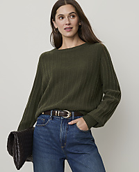 A woman in a green ribbed sweater and blue jeans holds a dark clutch in front of a plain background. carousel Product Image 1