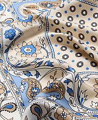A close-up of a beige silk scarf with a blue and brown paisley and floral pattern in the foreground, set against a background of similar patterned fabric. carousel Product Image 2