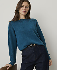 A woman in a teal sweater and dark jeans leans against a chair in front of a plain background. carousel Product Image 1