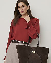 A woman in a red blouse and beige skirt holds a brown suede tote bag in front of a plain background. carousel Product Image 1