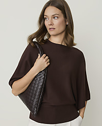 A woman in a brown sweater and white pants holds a woven brown purse against a plain background. carousel Product Image 1