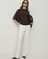 A woman in a brown sweater and white pants stands against a plain background, holding a woven bag. carousel Product Image 3