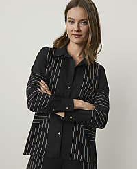 A woman in a black shirt with white geometric patterns stands with her arms crossed against a plain, light gray background.