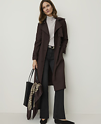 A woman in a dark brown trench coat and black flared jeans stands against a plain white background, holding a black tote bag with a patterned scarf. carousel Product Image 3