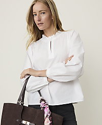 A woman in a white ruffled blouse holds a brown suede tote bag with a floral scarf in front of a plain, light-colored background. carousel Product Image 1