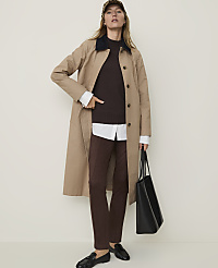 A woman in a trench coat and brown pants stands against a plain, light-colored background, holding a black tote bag. carousel Product Image 4