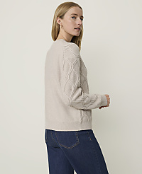 A woman in a beige cable-knit sweater and blue jeans stands against a plain, light-colored background. carousel Product Image 2