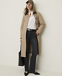 A woman in a tan trench coat, white blouse, dark jeans, and a leopard print cap stands against a plain, light-colored wall, holding a black tote bag. carousel Product Image 3