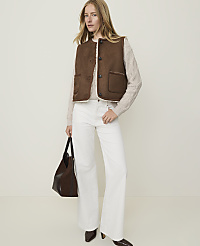 A woman in a brown vest and white pants stands against a plain, light background, holding a dark brown purse. carousel Product Image 3