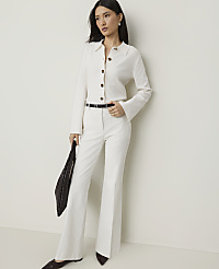 A woman in a white suit and dark shoes stands against a plain, light-colored wall, holding a dark woven bag. carousel Product Image 1
