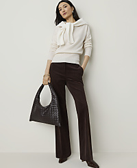 A woman in a cream sweater and brown corduroy pants stands with a dark woven bag against a plain white background.