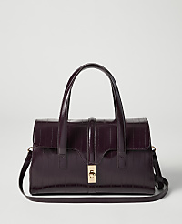 A deep purple, croc-embossed handbag with a gold clasp and top handles sits in the foreground against a plain, light gray background. carousel Product Image 1