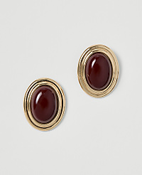 A pair of oval, gold-toned earrings with deep red cabochon stones are displayed against a plain, light gray background. carousel Product Image 1