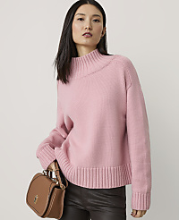 A woman in a pink sweater and dark pants holds a brown purse in front of a plain background. carousel Product Image 1